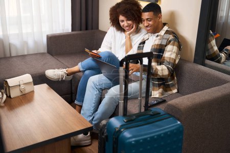 Téléchargez les photos : Les jeunes mariés en voyage sont assis sur un canapé dans une chambre d'hôtel avec des passeports et un ordinateur portable, les valises sont debout à proximité - en image libre de droit