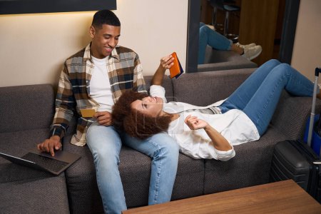 Téléchargez les photos : Femme bouclée allongée sur les genoux de son petit ami, couple relaxant sur le canapé dans le salon de la chambre d'hôtel - en image libre de droit