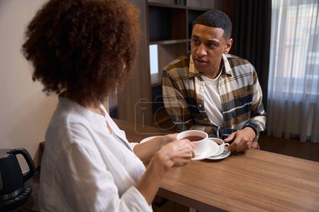Photo for Traveling multiracia spouses talking over morning coffee in hotel room - Royalty Free Image