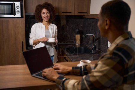 Téléchargez les photos : Couple multiracial bavardant dans la cuisine de la chambre d'hôtel pour le café du matin, l'homme a un ordinateur portable - en image libre de droit