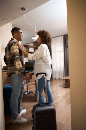 Téléchargez les photos : Couple multiracial satisfait se tient dans une chambre d'hôtel, deux valises à proximité - en image libre de droit