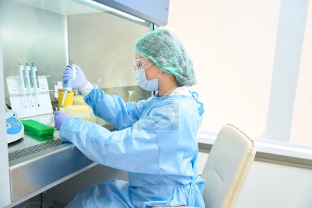 Laborantin mit Brille und Handschuhen sammelt Biomaterial mit einer speziellen Pipette, sie arbeitet unter sterilen Bedingungen