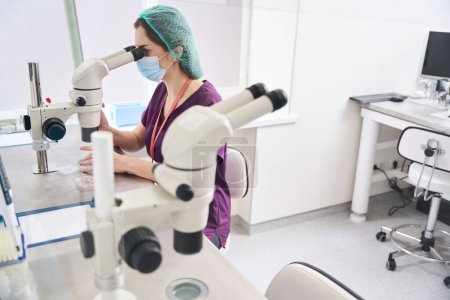 Photo for Woman laboratory assistant looks into the eyepiece of powerful microscope, at her workplace there is modern equipment for vitrification procedure - Royalty Free Image
