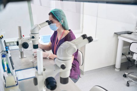 Foto de Trabajador médico en mono y máscara protectora mira en el ocular de un microscopio, el equipo para el procedimiento de vitrificación está alrededor - Imagen libre de derechos