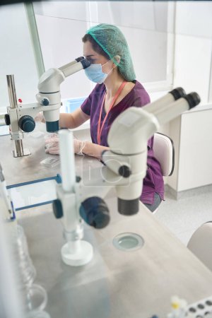 Foto de Mujer en mono y máscara protectora mira en el ocular de un microscopio de gran alcance, alrededor de equipos modernos para el procedimiento de vitrificación - Imagen libre de derechos