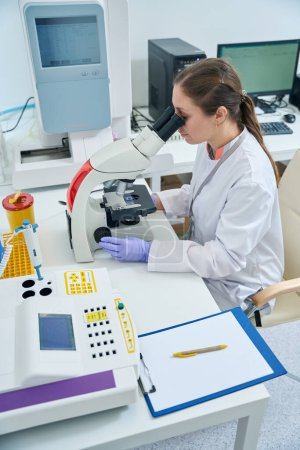 Photo for Hematologist examines a biomaterial sample through a microscope, a woman in protective gloves and overalls - Royalty Free Image
