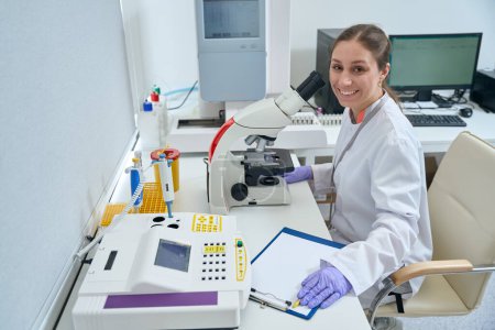Foto de Mujer sonriente se sienta en un lugar de trabajo en un laboratorio frente a un microscopio, entre los equipos de diagnóstico modernos - Imagen libre de derechos