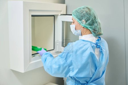 Photo for Virologist laboratory assistant transfers images of viruses from a sterile room to non-sterile one, woman in protective mask and overalls - Royalty Free Image