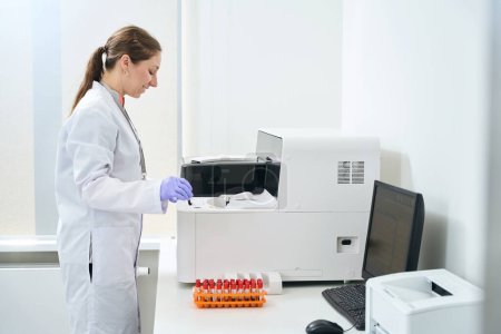 Foto de Asistente de laboratorio se encuentra cerca del analizador hematológico en la unidad de prueba del laboratorio moderno, una computadora y una impresora están en la mesa - Imagen libre de derechos