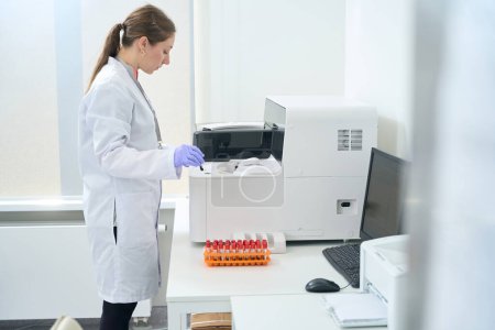 Photo for Laboratory assistant works with a hematological analyzer in a testing unit of modern laboratory, computer and printer are on table - Royalty Free Image