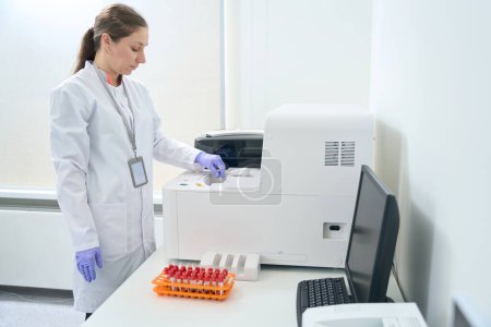 Téléchargez les photos : Hematologist laboratory assistant stands near an immunochemiluminescent analyzer in the testing unit of a modern laboratory - en image libre de droit