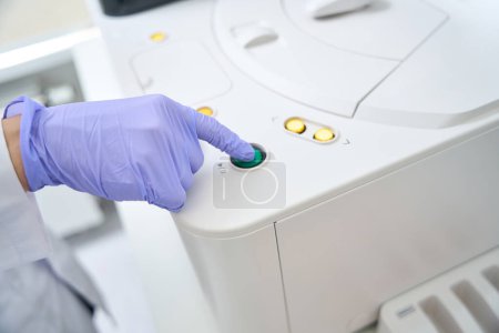 Téléchargez les photos : L'assistant de laboratoire appuie sur le bouton sur le corps de l'analyseur immunochimiluminescent, l'agent de santé dans des gants de protection - en image libre de droit