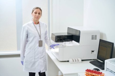 Téléchargez les photos : Une généticienne se tient dans l'unité de test, sa main repose sur un analyseur immunochimiluminescent - en image libre de droit