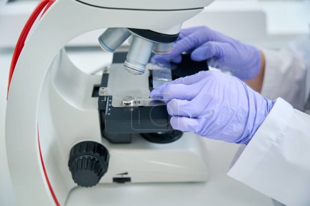 Foto de Empleado de laboratorio coloca un biomaterial para una prueba de ADN bajo un microscopio, un especialista trabaja en guantes de protección - Imagen libre de derechos