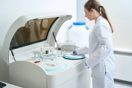 Foto de Mujer en overoles se coloca frente a un analizador bioquímico moderno, se utiliza para obtener muestras de hígado, riñón - Imagen libre de derechos