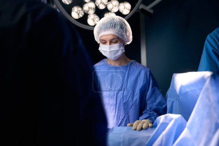 Téléchargez les photos : Une femme portant un masque de protection et un uniforme est debout dans la salle d'opération, à côté de ses collègues - en image libre de droit