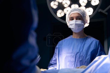 Téléchargez les photos : Femme dans un masque de protection et uniforme se tient à la table d'opération, à côté de ses collègues - en image libre de droit