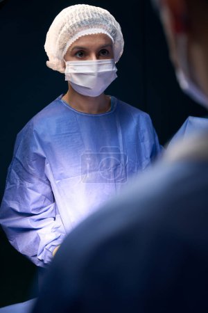 Foto de Mujer en uniforme quirúrgico estéril se encuentra bajo la luz de una lámpara, junto a su colega - Imagen libre de derechos