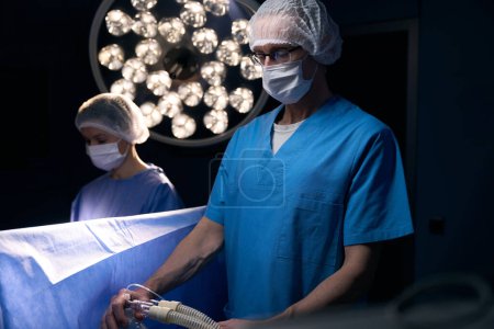 Foto de Anestesiólogo en mono quirúrgico está en la mesa de operaciones, un asistente trabaja cerca - Imagen libre de derechos