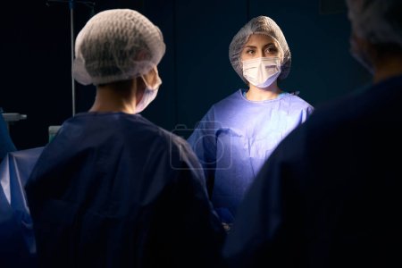 Téléchargez les photos : Groupe de femmes en uniformes chirurgicaux debout à la table d'opération, au centre est une femme dans un masque - en image libre de droit