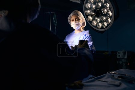 Foto de Una mujer con uniforme quirúrgico se para en la mesa de operaciones, le pasa el instrumento a su colega - Imagen libre de derechos