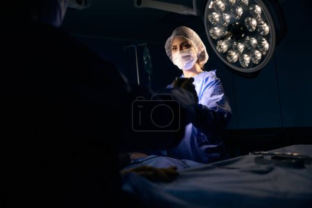 Foto de Mujer en una máscara protectora se para en la mesa de operaciones, ella pasa el instrumento a su colega - Imagen libre de derechos