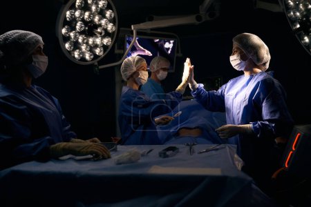 Foto de Colegas cirujanos dan cinco después de una operación exitosa, el paciente se acuesta en la mesa de operaciones - Imagen libre de derechos