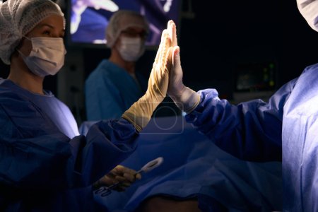 Foto de Médicos en el quirófano dan cinco altos después de una operación exitosa, un paciente se acuesta en la mesa de operaciones - Imagen libre de derechos