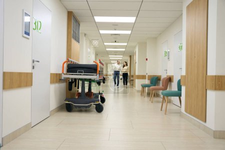 Téléchargez les photos : Femme aux cheveux foncés et homme d'âge moyen marchent le long du couloir de l'hôpital, au premier plan contre le mur est un lit mobile - en image libre de droit
