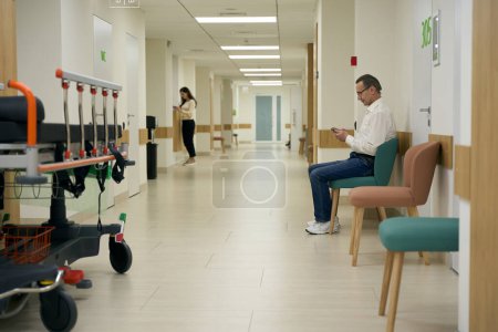 Foto de Hombre con gafas se sienta en el pasillo del hospital con un teléfono en las manos, una mujer está de pie en el fondo - Imagen libre de derechos
