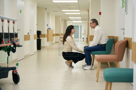 Téléchargez les photos : La gentille jeune femme réconforte un homme d'âge moyen dans un couloir d'hôpital, au premier plan se trouve un lit mobile - en image libre de droit