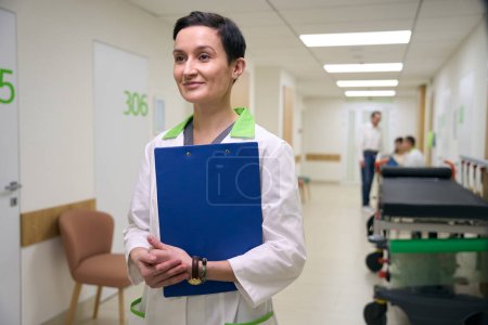 Foto de Mujer en puestos de bata médica con carpeta azul en el pasillo del centro médico, en el fondo la gente en ropa casual - Imagen libre de derechos