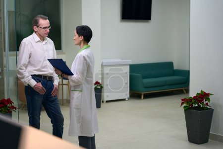 Téléchargez les photos : Une travailleuse de la santé communique avec un visiteur dans le hall de l'hôpital, la femme a un dossier bleu dans ses mains - en image libre de droit