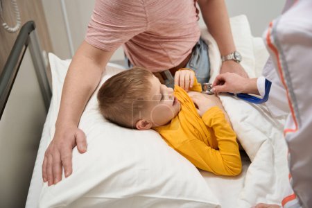 Téléchargez les photos : Portrait de vue latérale du petit garçon soigné est couché sur le lit médical tandis que le médecin examine les indicateurs du corps avec des équipements spéciaux à l'hôpital - en image libre de droit