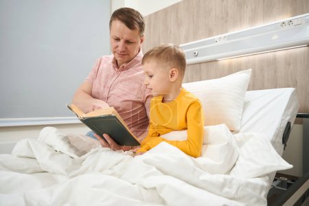 Foto de Cintura arriba retrato de hombre caucásico guapo está leyendo el libro para su pequeño hijo en el cuarto de los enfermos - Imagen libre de derechos
