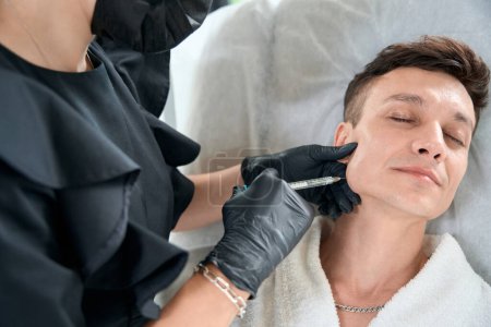 Téléchargez les photos : Cosmétologue professionnel dans des gants de protection tenant la seringue et faisant des injections de beauté pour le visage à l'homme - en image libre de droit