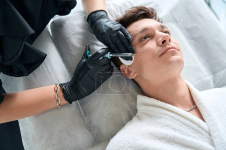 Téléchargez les photos : Médecin dans des gants de protection tenant la seringue et faisant des injections de beauté dans le visage à bel homme - en image libre de droit