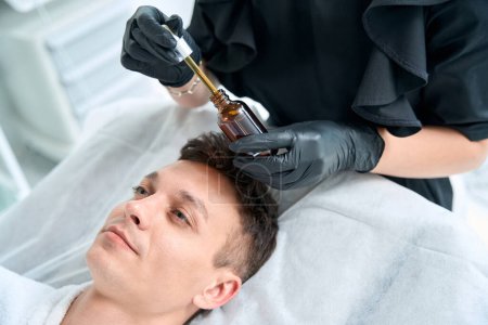 Téléchargez les photos : Cosmetologue dans des gants de protection tenant l'huile pour la peau et appliquer le produit sur le visage du patient dans le salon - en image libre de droit
