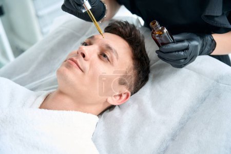 Téléchargez les photos : Dermatologue dans des gants de protection tenant l'huile pour la peau et appliquer le produit sur le visage du patient dans le salon - en image libre de droit