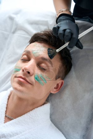 Téléchargez les photos : Cosmetologue dans des gants de protection tenant un bocal avec masque pour la peau et appliquer le produit avec brosse sur le visage du patient - en image libre de droit