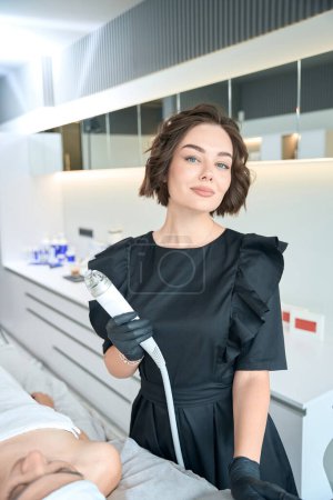 Photo for Cosmetologist in protective gloves standing near adult female and holding microneedle rf-lifting instrument in beauty salon - Royalty Free Image
