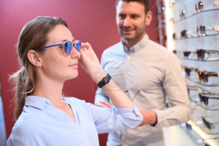 Téléchargez les photos : Gros plan vue de côté portrait de charmante femme caucasienne essaie les lunettes de soleil de luxe avec l'homme dans le magasin opticien - en image libre de droit
