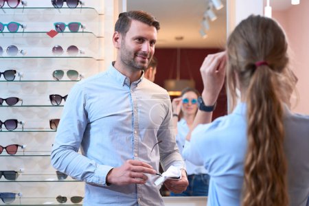 Photo for Close up portrait of handsome Caucasian man is helping to the lady with chose of bright luxury sunglasses in optician store - Royalty Free Image