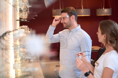 Photo for Side view portrait of handsome smiling man is wearing glasses while looking to the reflection in the luxury optician shop - Royalty Free Image