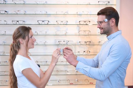 Photo for Waist up side view portrait of handsome smiling man is trying the glasses and asking the shop assistant about optical fashion in the store - Royalty Free Image