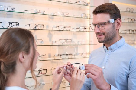 Photo for Close up portrait of handsome Caucasian man is trying the eyeglasses while female shop assistant is consulting client in the optical store - Royalty Free Image