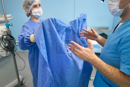 Téléchargez les photos : Assistant dans la salle préopératoire aide le chirurgien à mettre une robe, les médecins dans des masques de protection - en image libre de droit