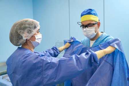 Téléchargez les photos : Infirmière dans la salle préopératoire aide le chirurgien à mettre une robe stérile, les médecins dans des masques de protection - en image libre de droit