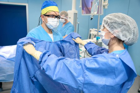 Téléchargez les photos : Infirmière aide le médecin à mettre une robe stérile, le chirurgien a une lampe spéciale sur son front - en image libre de droit