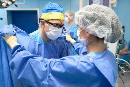 Téléchargez les photos : Processus de préparation à une opération chirurgicale, l'infirmière aide le médecin à revêtir une robe stérile - en image libre de droit